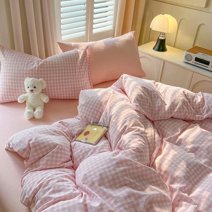 a teddy bear sitting on top of a bed with pink comforter and pillows in front of a window