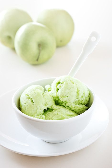 a bowl filled with green ice cream next to three apples on a white tablecloth