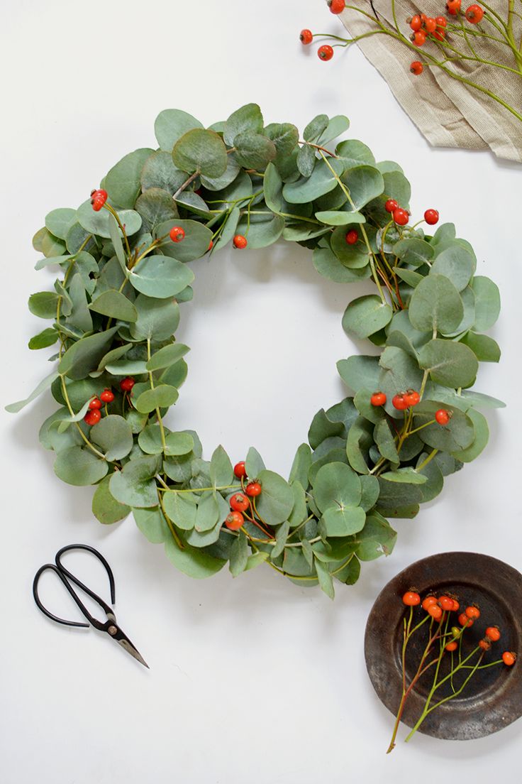 a wreath with red berries and greenery next to scissors on a white table top