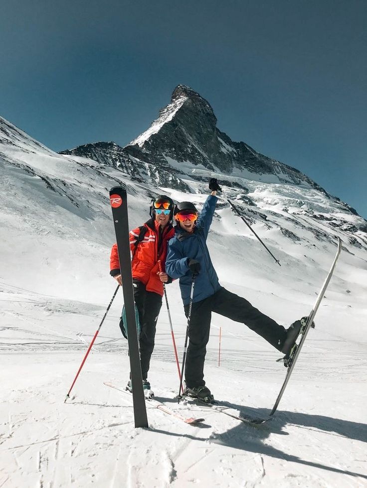 two skiers pose for the camera in front of a snow covered mountain with their skis on