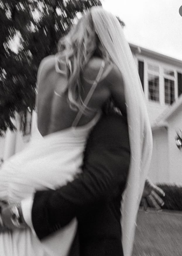 black and white photograph of a bride carrying her groom on his back in front of a house