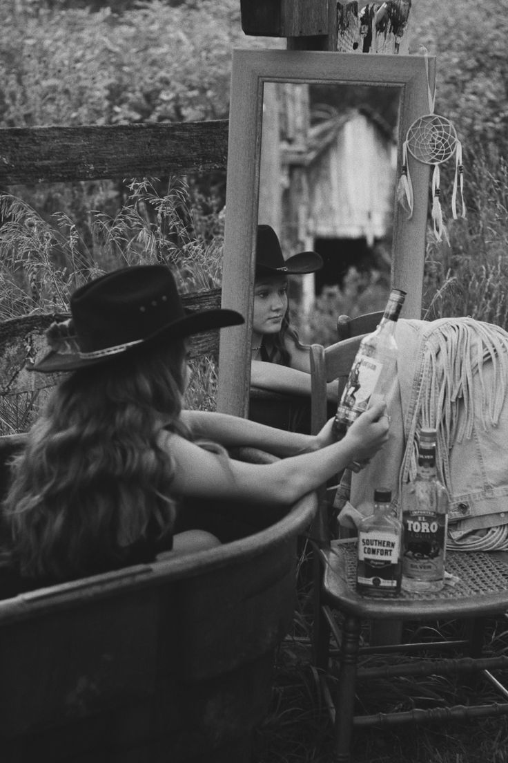 a woman sitting in a bathtub with a cowboy hat on, looking at herself in the mirror