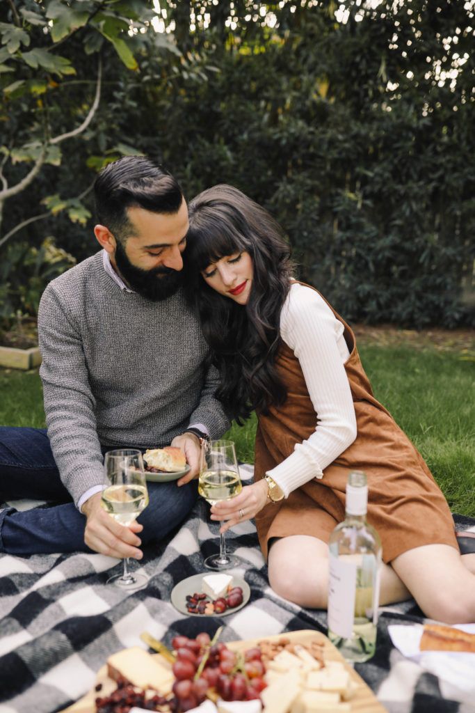 a man and woman sitting on the grass with wine glasses in their hands while they are holding plates of food