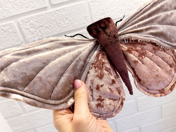 a person holding a stuffed butterfly in front of a white brick wall with brown spots on it's wings