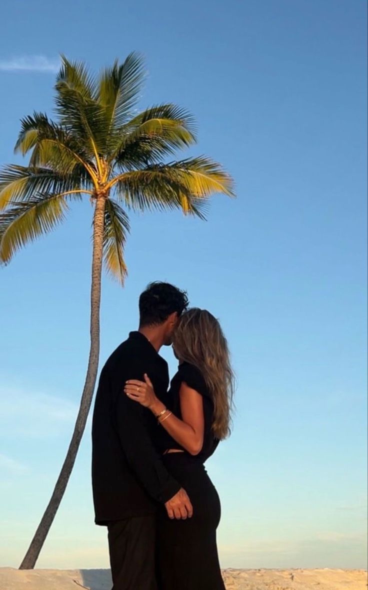 a man and woman standing next to each other on a beach under a palm tree