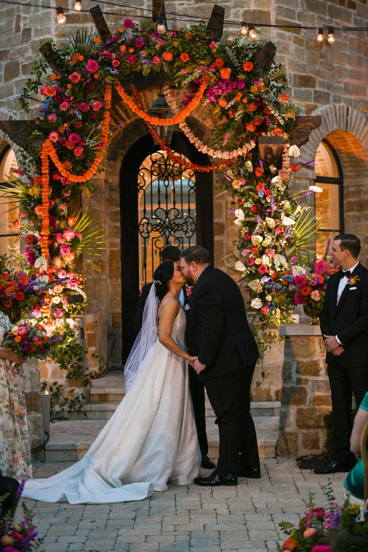 a bride and groom kiss in front of an archway decorated with colorful florals at their wedding