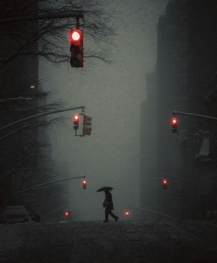 a person with an umbrella walking across a street in the snow at night under traffic lights