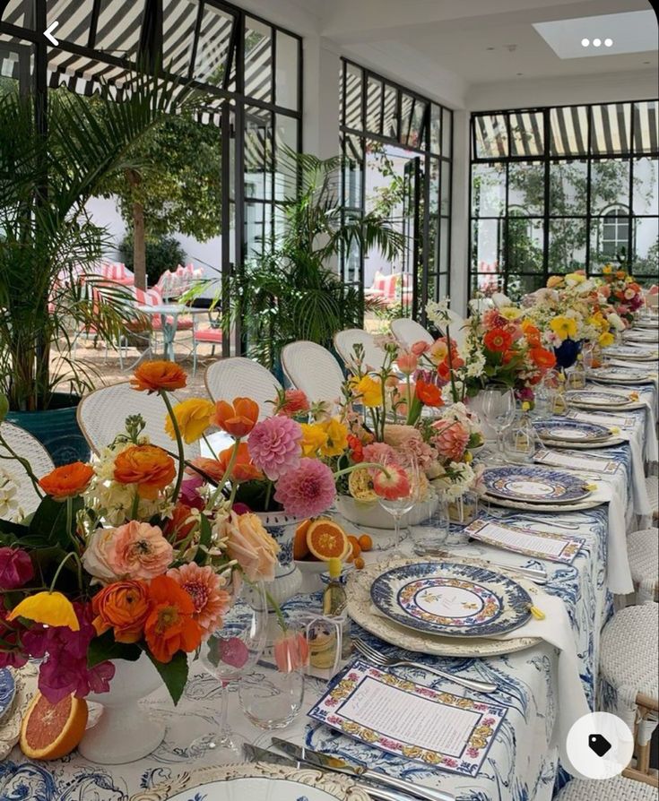 a long table is set with plates and vases filled with colorful flowers in front of large windows