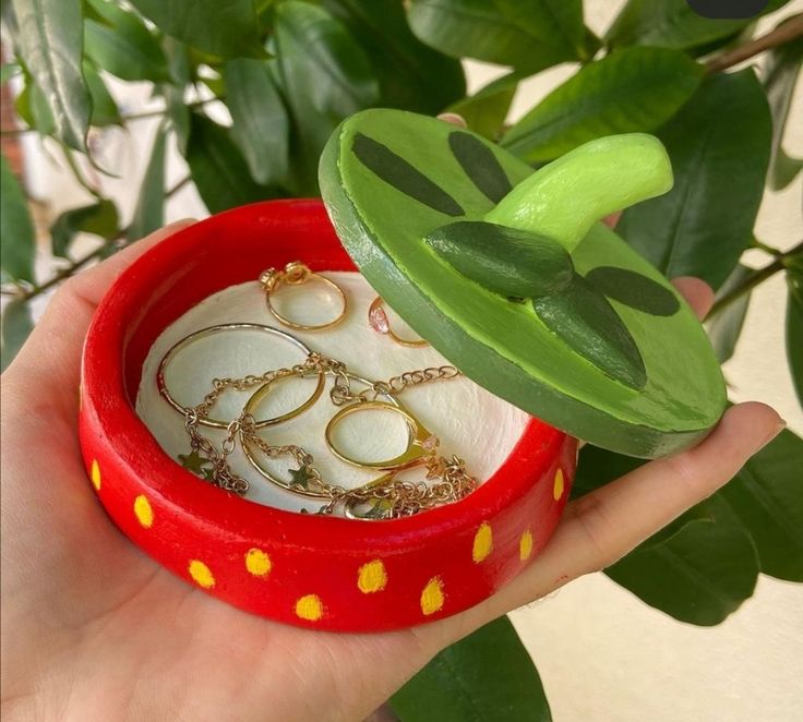 a hand holding a red ring box with gold rings in it and a green plant behind it