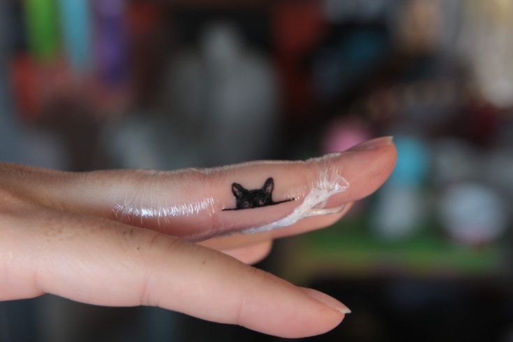 a tiny black cat tattoo on the tip of a finger, by someone's fingers