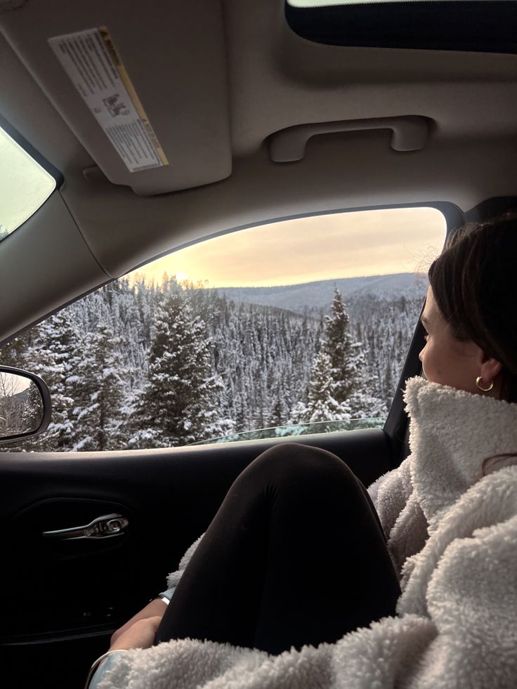 a woman sitting in the back seat of a car looking out at snow covered trees