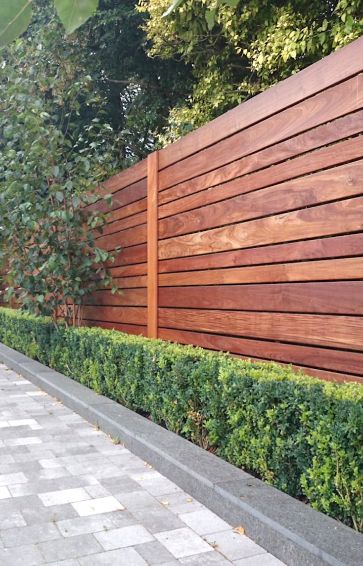 a wooden fence next to a sidewalk and some bushes on the side of a building