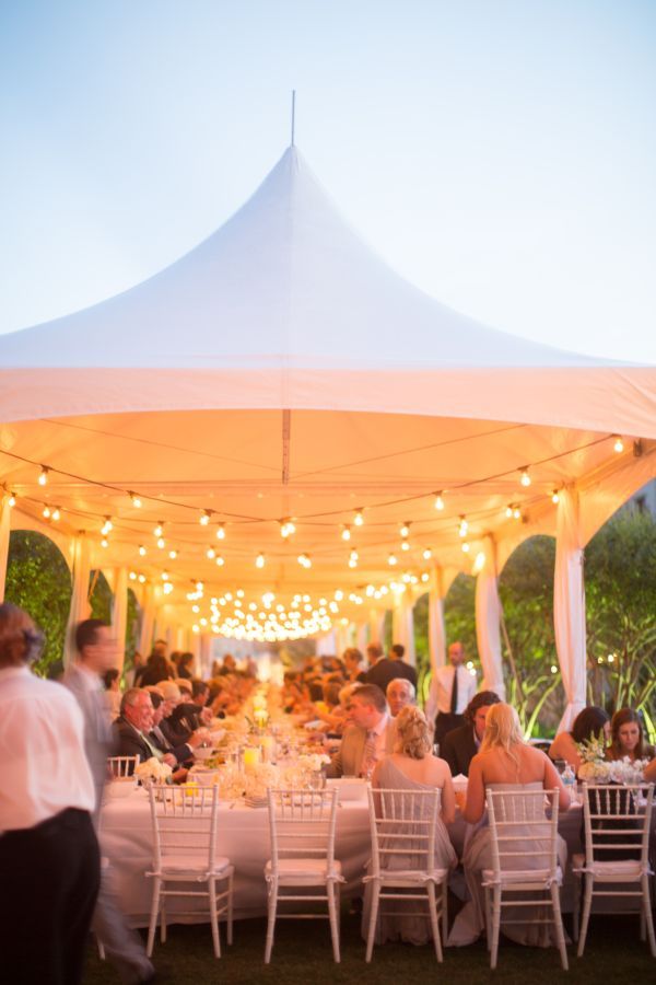 a group of people sitting at tables under a white tent with lights on the ceiling