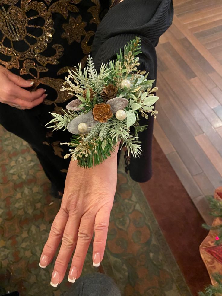 a woman's hand holding a bouquet of greenery and pine cones on her wrist