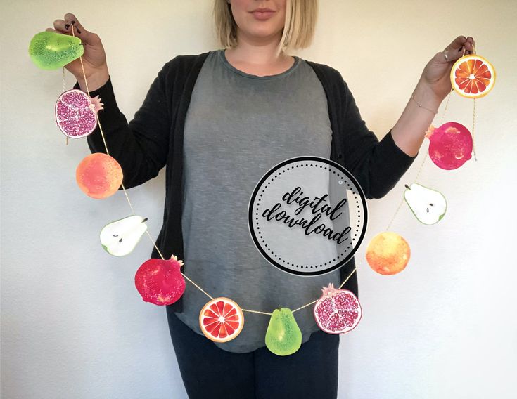 a woman standing in front of a white wall holding fruit garlands with the words happy holidays written on them