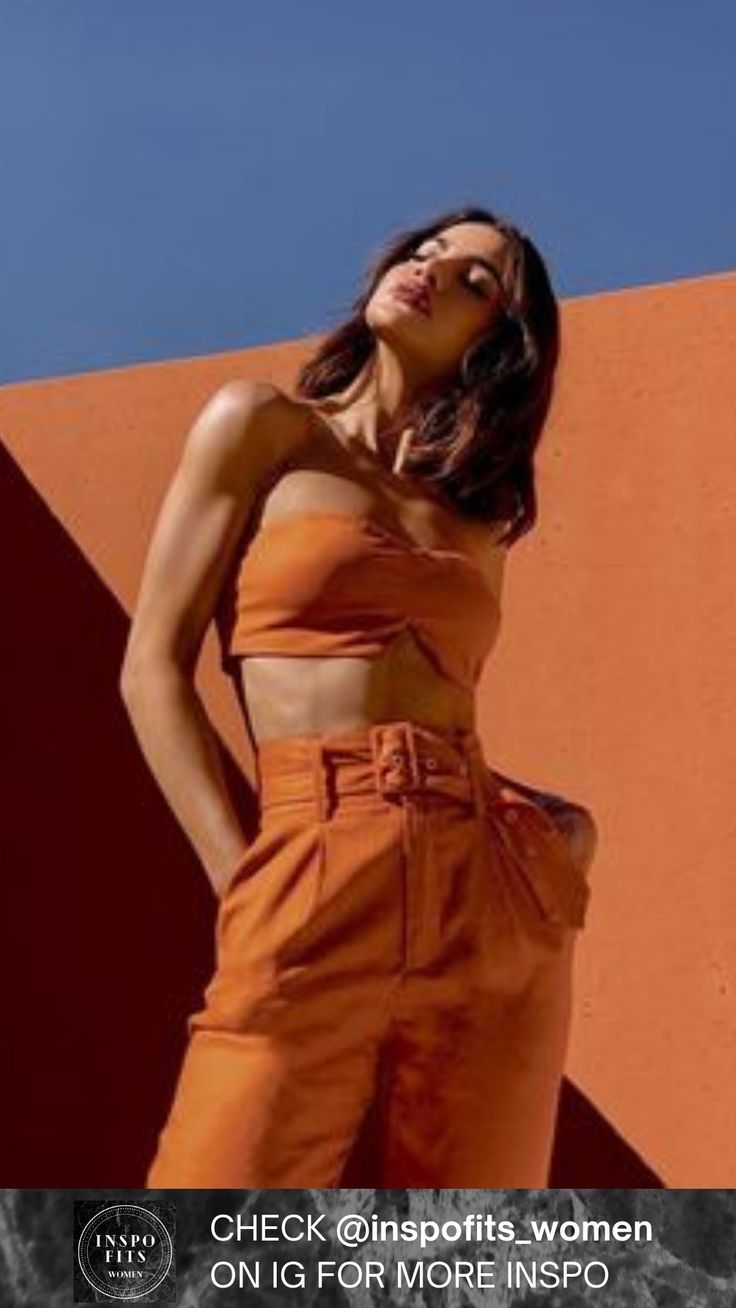 a woman in an orange top and pants standing next to a wall with her hands on her hips