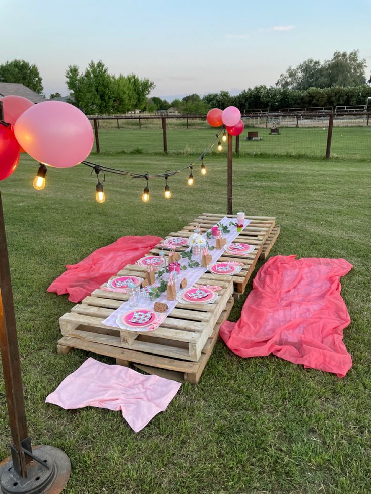 a picnic table set up in the middle of a field with balloons and pink napkins