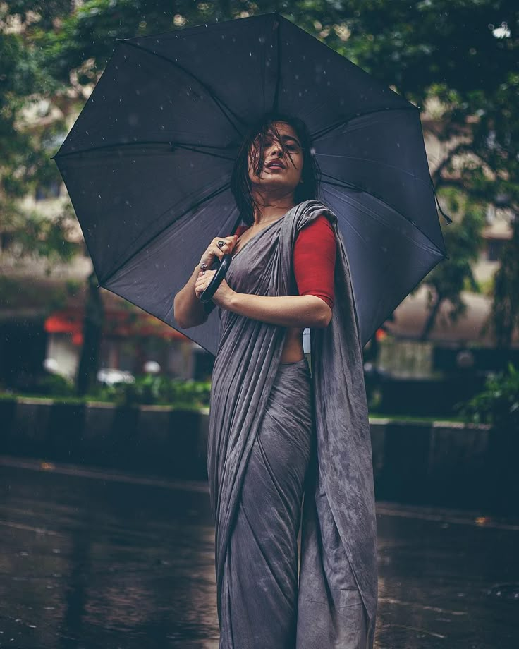 a woman standing in the rain holding an umbrella and wearing a gray sari with red lining