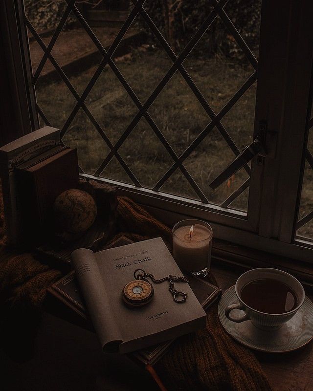 a cup of tea, book and pocket watch sitting on a table next to a window