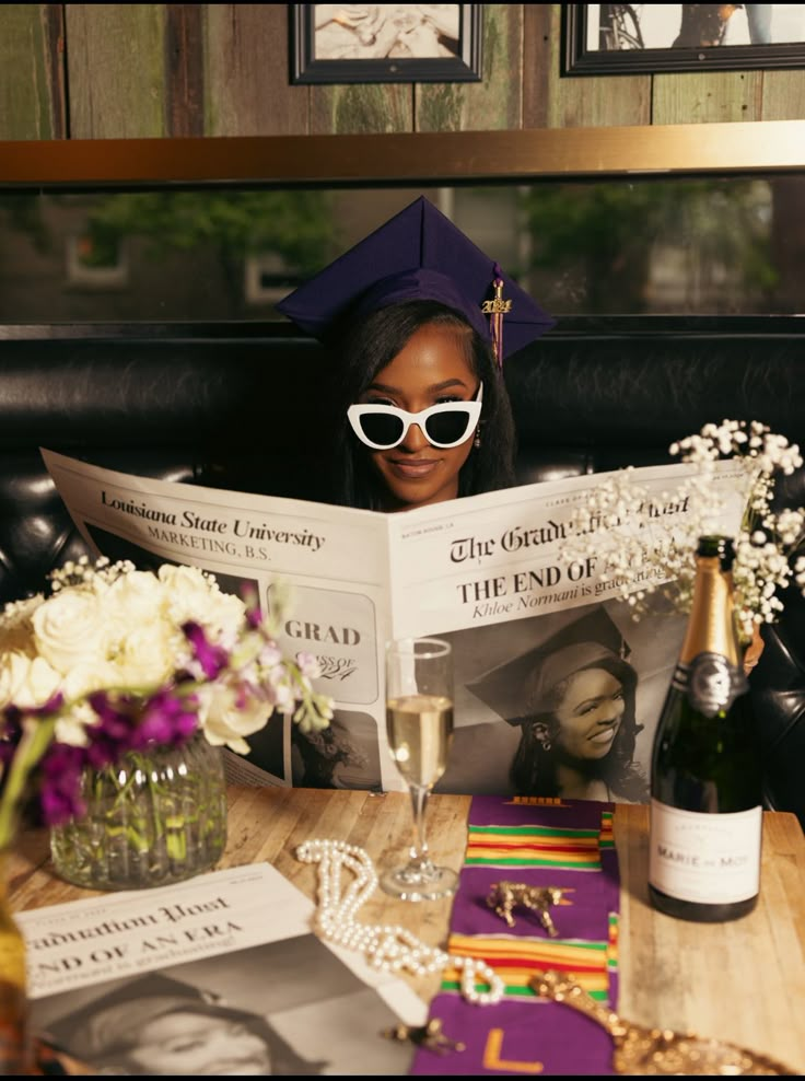 a woman reading a magazine while sitting at a table with wine and flowers in front of her