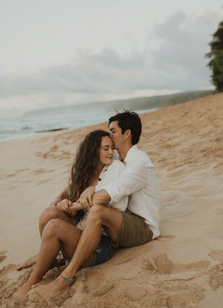 a man and woman are sitting on the sand at the beach, hugging each other