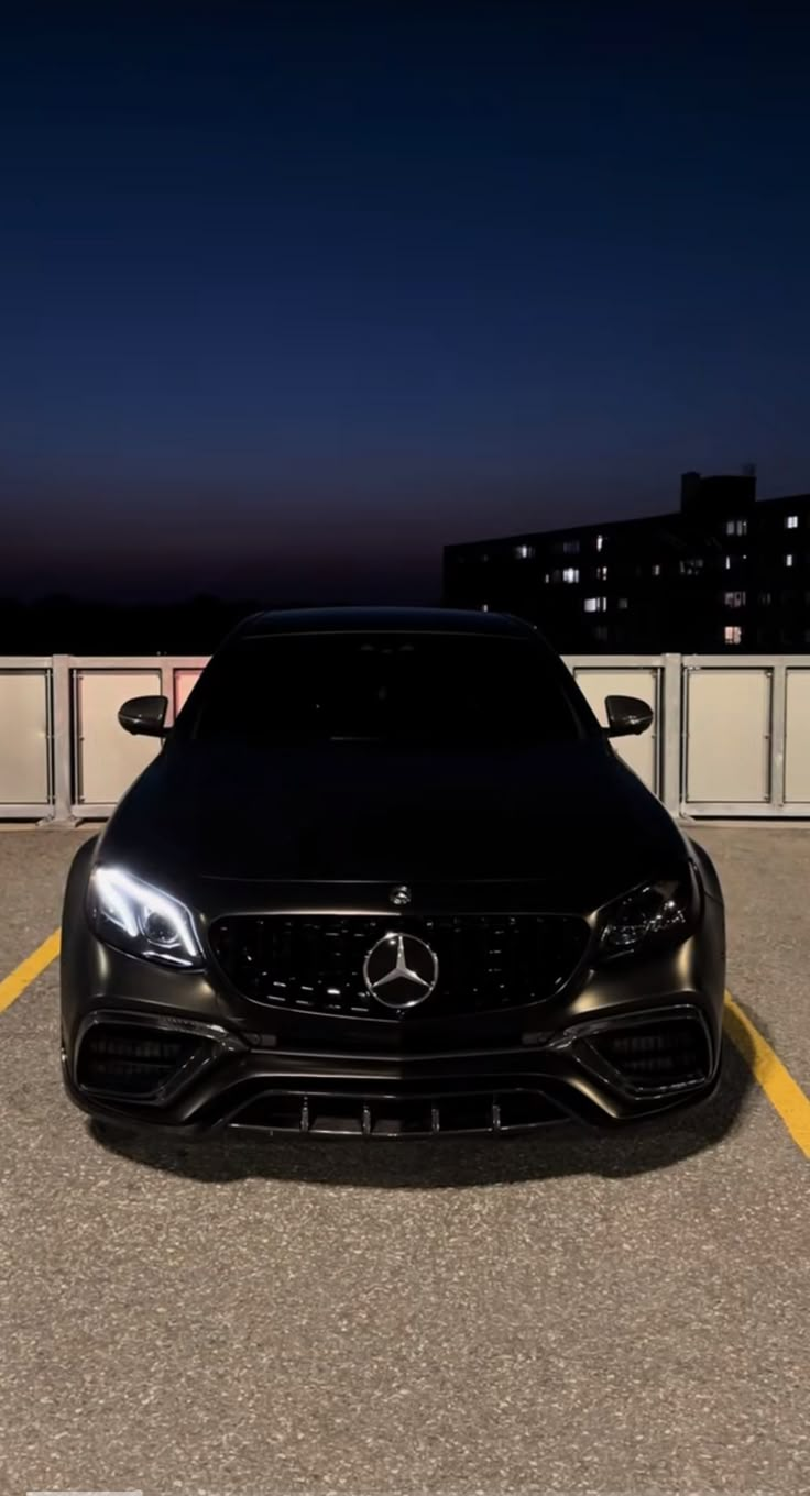 the front end of a black car parked in a parking lot at night with buildings in the background