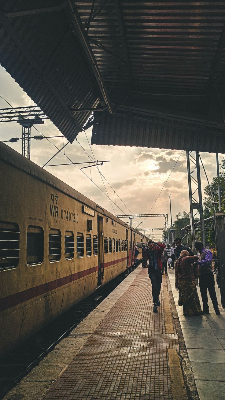 people are walking on the sidewalk next to a train at an outdoor station as the sun sets