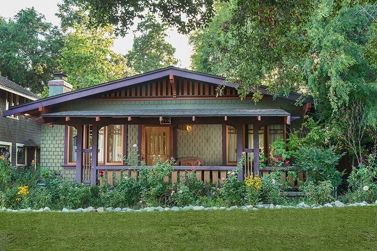 a small wooden house surrounded by trees and flowers in front of some bushes on the lawn