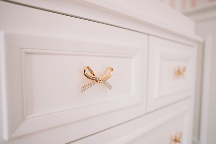 a close up of a white dresser with gold handles and bows on the knobs