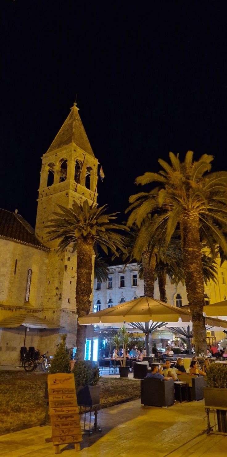 palm trees in front of an old building at night with people sitting on benches under umbrellas