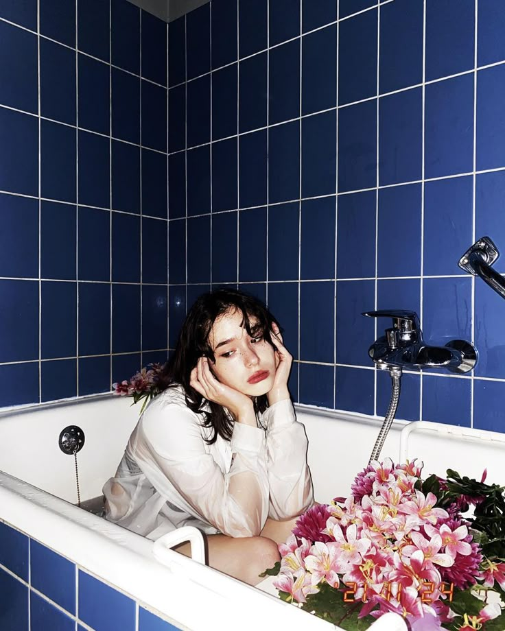 a woman sitting in a bathtub with flowers on the side and blue tiled walls