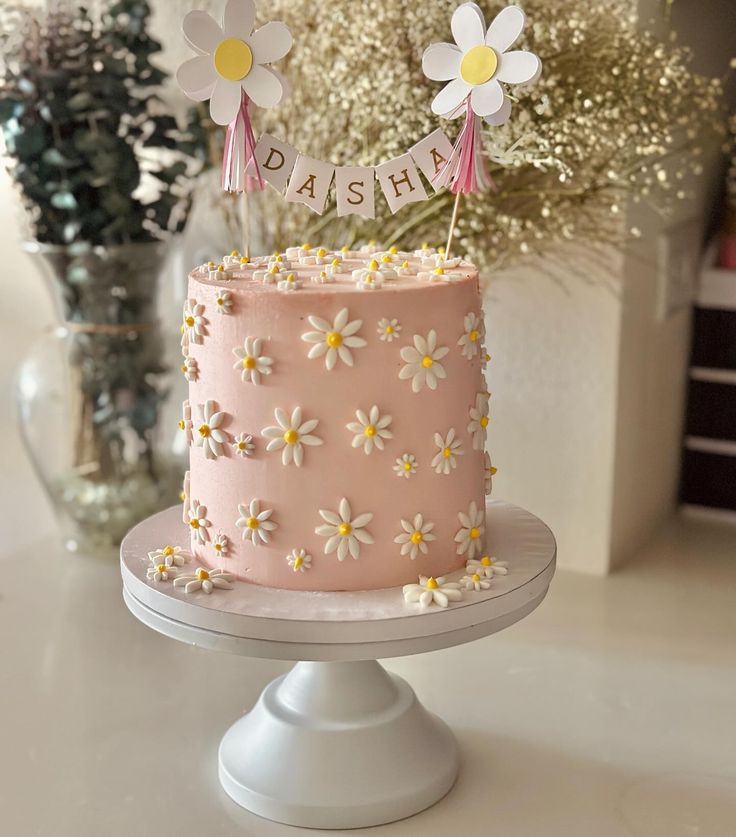 a pink cake with daisies on top sitting on a table next to some flowers