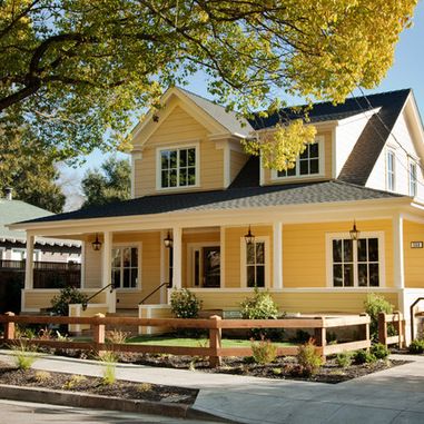 a yellow house with black roof and white trim on the front porch is surrounded by trees