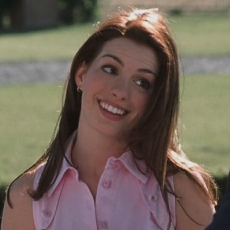 a woman with long brown hair wearing a pink shirt smiles at the camera while standing in front of a grassy area