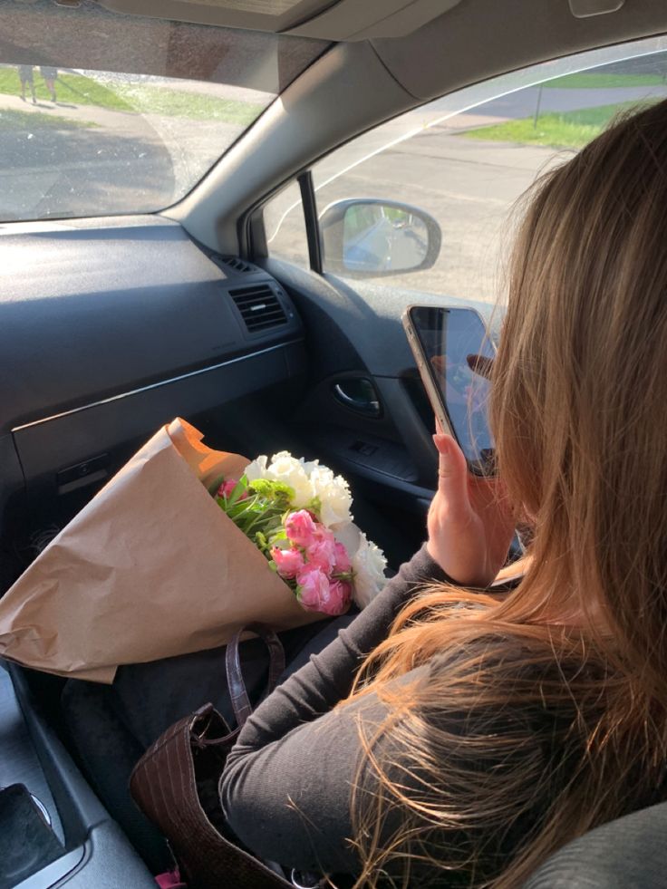 a woman sitting in the back seat of a car holding a cell phone and flowers