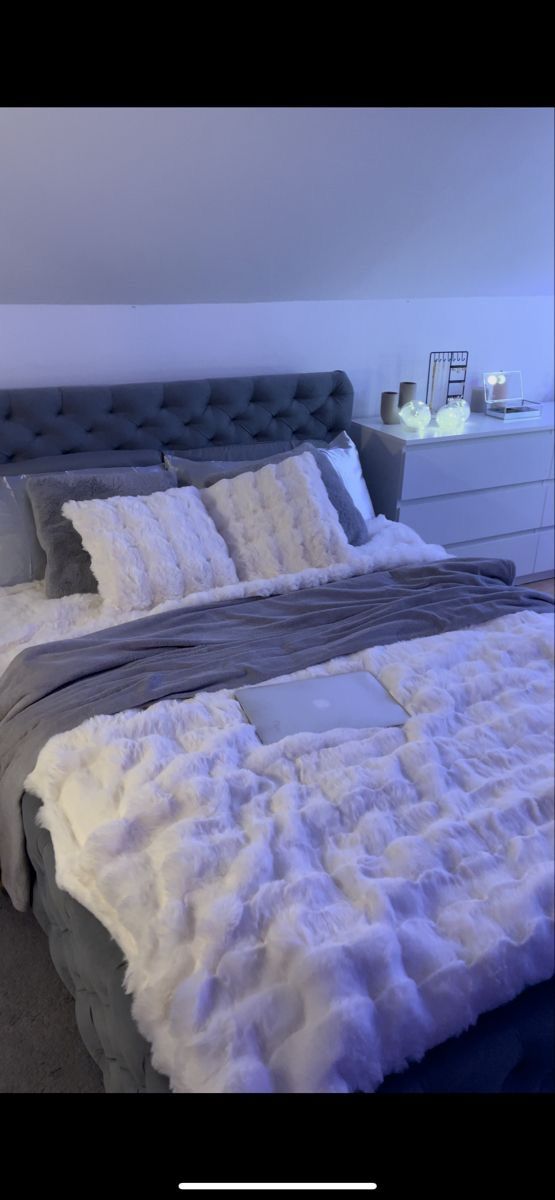 the bed is made with white fur and grey linens, along with two night stands on either side