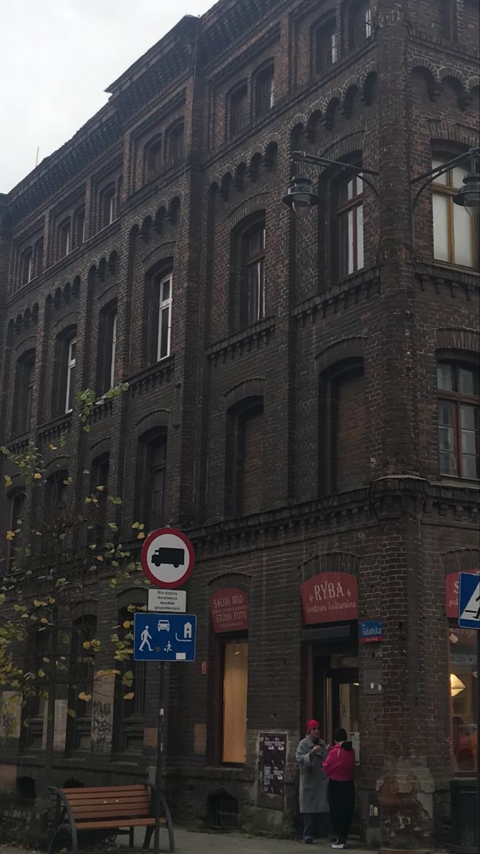 an old brick building with many windows and people standing in front of it on the sidewalk