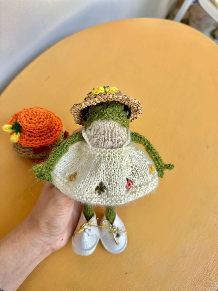 a hand holding a small stuffed animal on top of a wooden table next to a knitted hat