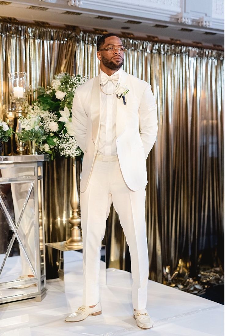 a man in a white tuxedo standing next to another man wearing a suit