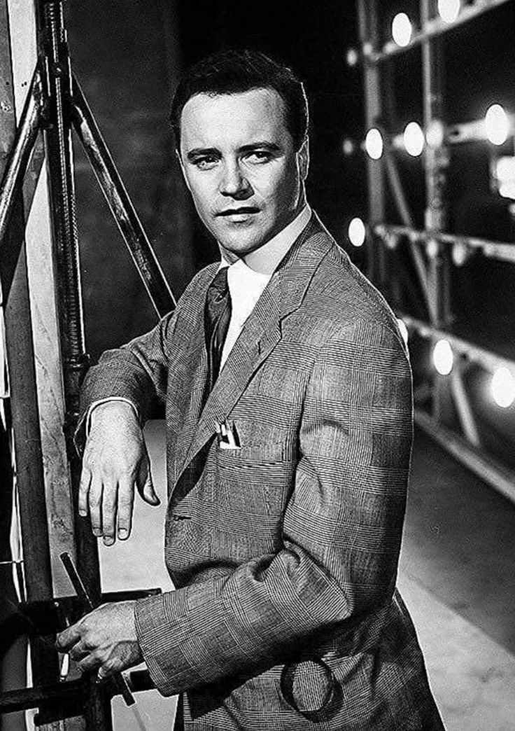 a black and white photo of a man in a suit leaning against a fence with his hand on the railing