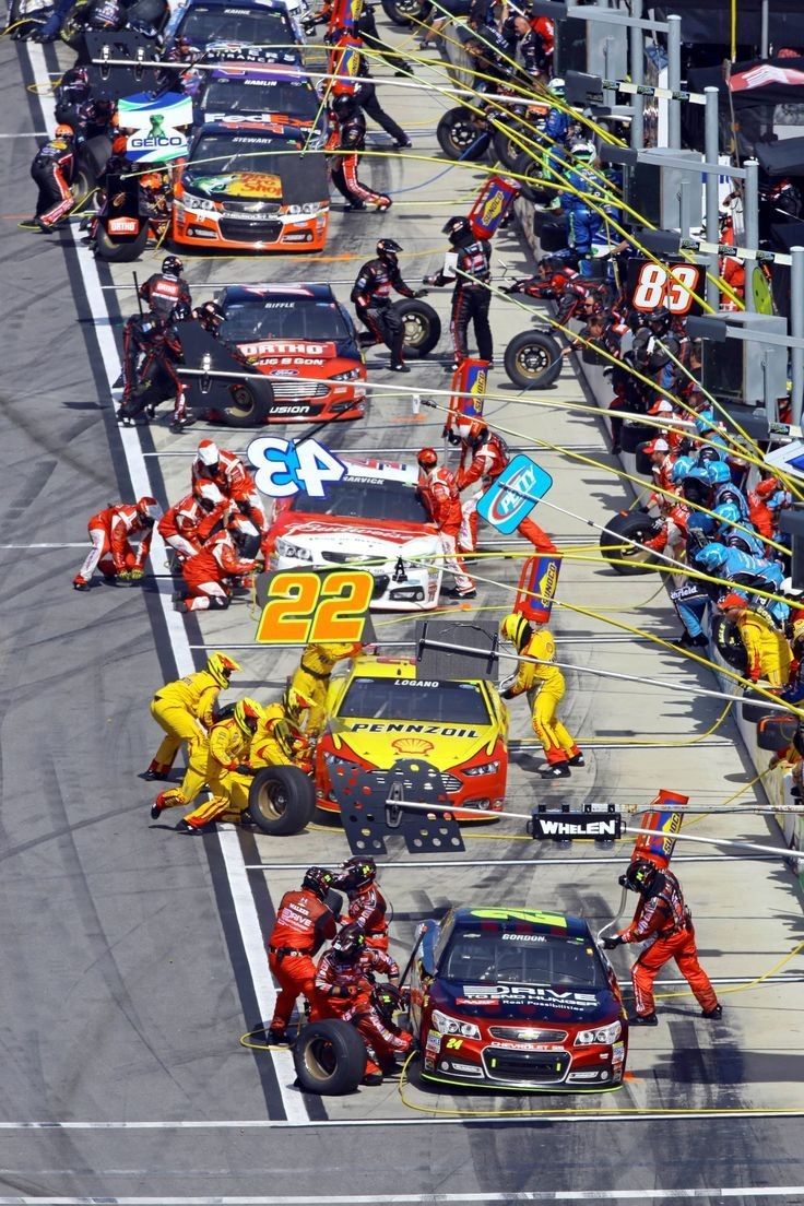 a group of people in red and yellow suits around cars on a race track with one person standing next to the car