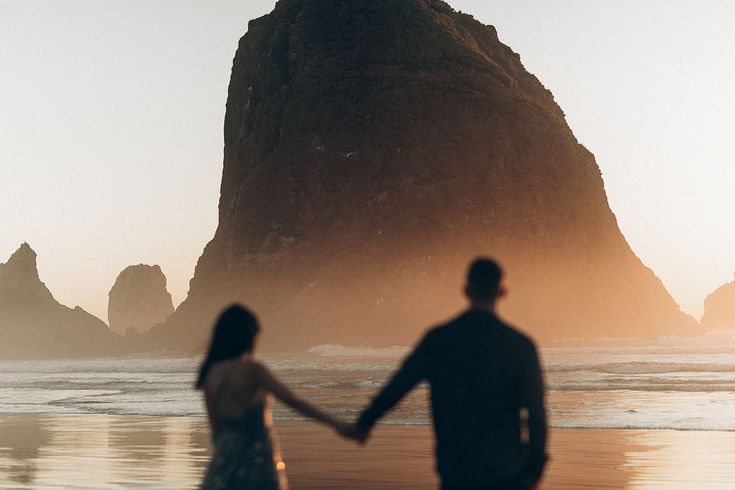 a man and woman holding hands walking on the beach next to an ocean with a rock formation in the background