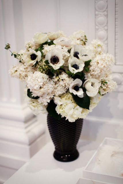 a black vase filled with white flowers sitting on top of a table next to a wall