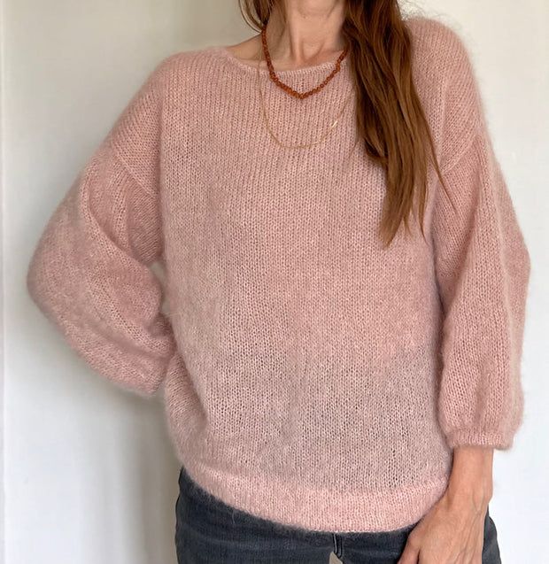 a woman standing in front of a white wall wearing a pink sweater