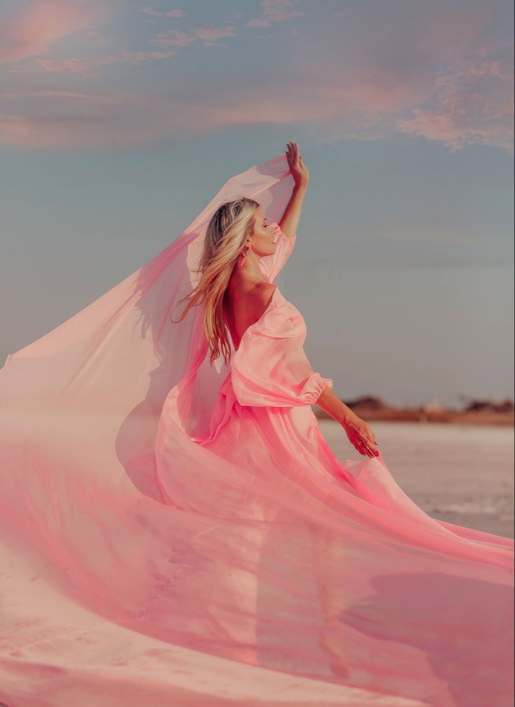 a woman in a long pink dress is standing on the beach with her arms outstretched