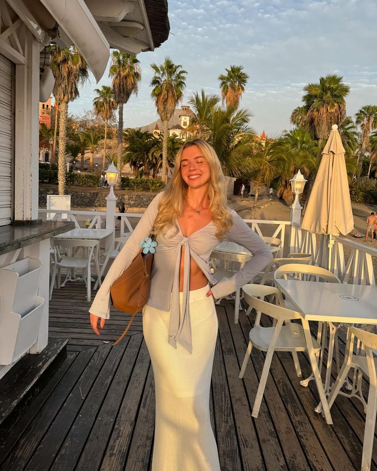 a woman standing on a wooden deck with palm trees in the background and white chairs around her