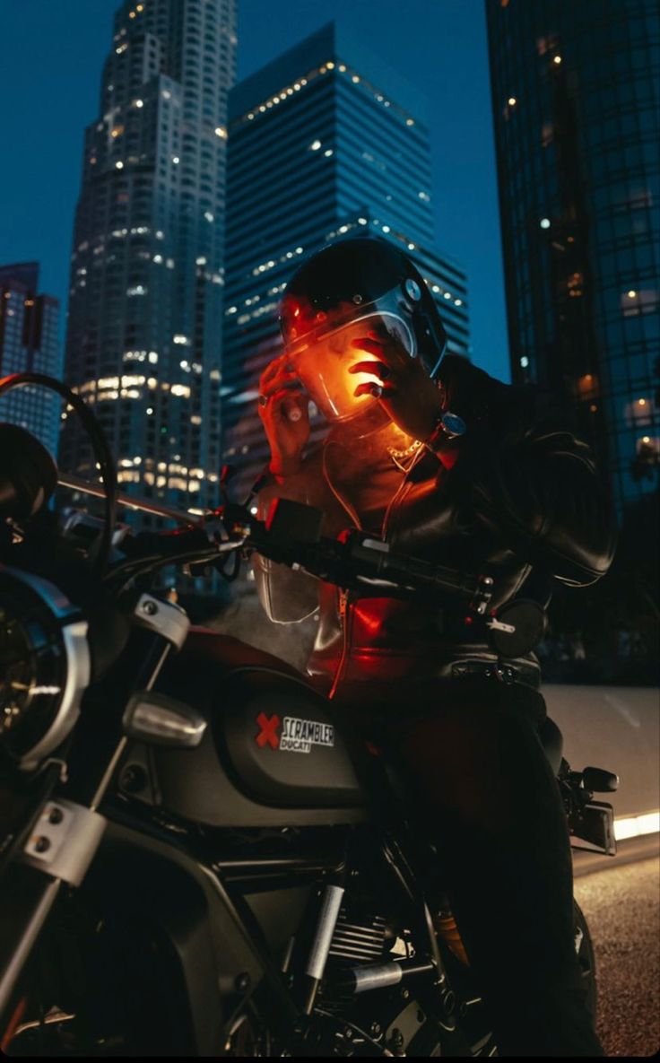 a man sitting on top of a motorcycle in front of a city skyline at night