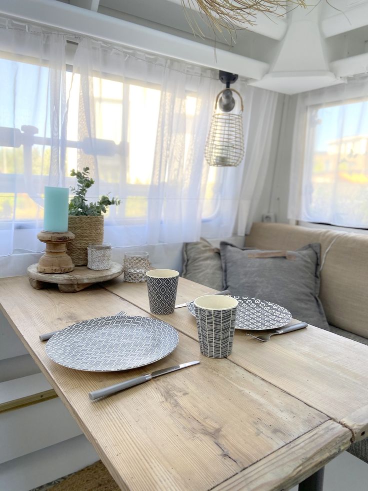 a wooden table with plates and cups on it in front of a couch next to a window