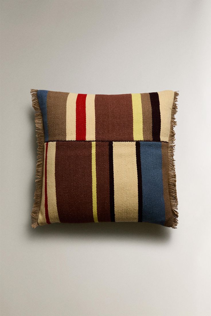 a multicolored striped pillow with fringes on the front and back, against a white wall