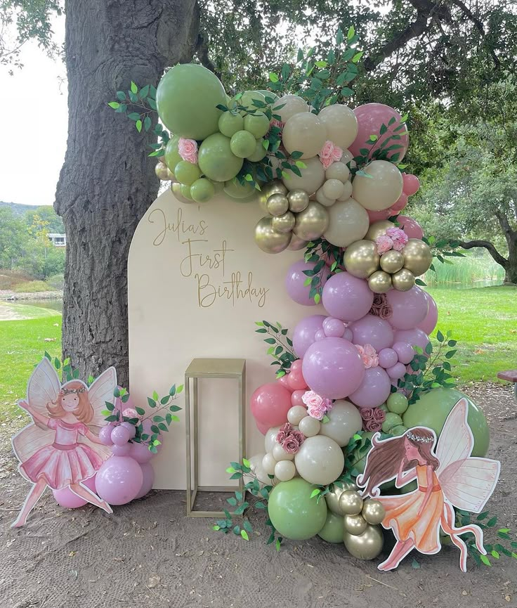 a birthday arch with balloons and fairy figurines on the ground next to a tree